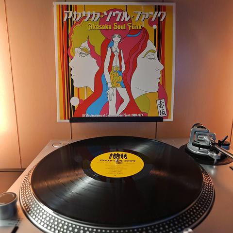 A vinyl record sits on a turntable. Behind the turntable, a vinyl album outer sleeve is displayed. The front cover shows artwork of a woman wearing a 70s style outfit and she has an afro. There is also a woman's face on the person's left, and man's face on the right.