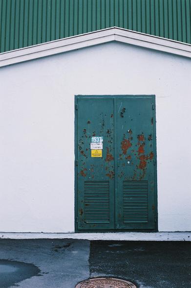 Photo of a green double door to an electric transformer room attached to a building. Around the industrial-looking labels saying ‘danger, high voltage’, the paint is flaking off the door, revealing rusty brown scars underneath. The doors are sitting on a white concrete wall for the transformer room, but in the background, the wall of the taller main building matches the green of the doors.