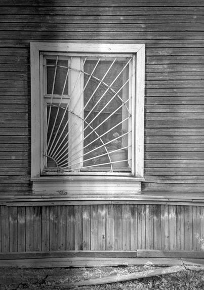 Monochrome photo shows a close-up of a window of an old wooden house. There is a metal grate in the form of a spider's web on the window.