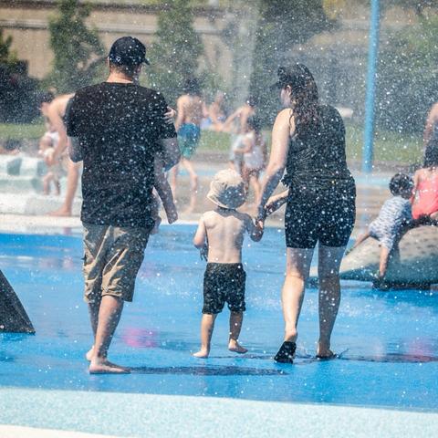 In the image, a family is walking along a path near a fountain, where water is spraying in all directions. The parents are holding hands with their child, who is dressed in a black tank top and shorts. There are other people in the background, also enjoying the park and walking along the path.