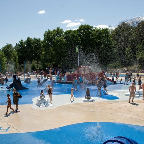 A blue water park is bustling with activity on a sunny day. The park features a splash pad where people are playing, and the park is filled with numerous visitors who are engaged in various water activities such as splashing in the water or sitting on large inflatable objects. The park is surrounded by trees, and the sky above is clear and blue. The visitors are seen enjoying their time at the water park and socializing with one another.