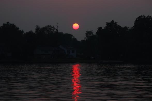 Sunset photo of a red and orange sun in a dark grey sky over a tree-lined lake with the red light of the sun reflecting in the surface of the water.