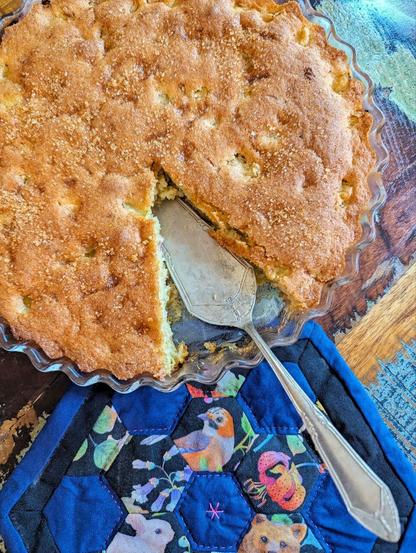 Dunkler Tisch. In der oberen Bildhälfte eine gläserne Tarteform mit gewelltem Rand. Darin braun gebackener Kuchen. eine Kuchenecke fehlt, ein Tortenheber liegt an dieser Stelle. In der unteren Bildhälfte ist ein kleiner Mini-Quilt in Hexagonform zu sehen. In Dunkelblau und schwarz mit pinkem Quiltgarn. die Patchworkstücke haben teils Tier- und Pflanzenmotive.