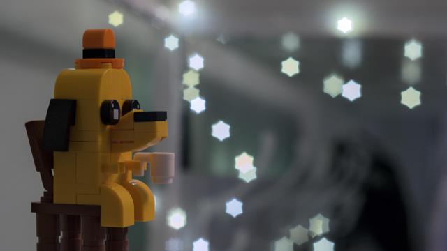 A yellow "this is fine" dog made from lego parts, with a cup in its hand, sitting in front of a blurry grey background with a number of star-shaped bokeh lights