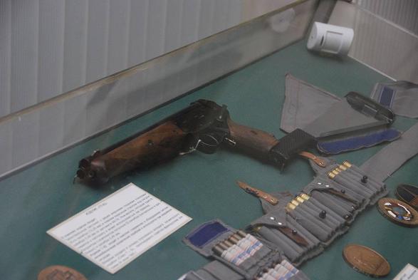 Ein in einem Glasdisplay befindliches Handfeuerwaffe, die TP-82, mit einem braunen Griff. Um die Waffe herum liegen einige Munitionsteile und weitere Gegenstände in einem glatten, grünen Untergrund. Ein erklärender Text befindet sich unter der Waffe.