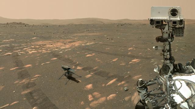 A wide shot showing part of Perseverance, and Ingenuity on flat reddish-brown gravel with a largely flat landscape behind them, and a small border of hills. The visible part of Perseverance is most wires and metal parts clustered together to house some small instruments. Ingenuity is a metal box, topped with two rotary blades and a solar panel, standing on four thin metal legs.