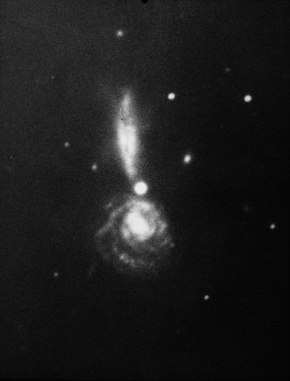 A grainy, black and white image of a pair of spiral galaxies. The lower galaxy is seen face-on. It has a bright center that transitions into a series of loosely-wound spiral arms, looking something like a cinnamon roll. The arms are dotted with star clusters . The upper galaxy is seen edge on. It has a somewhat puffy disk, seen as a wide, vertical line. Together, the two galaxies look like an exclamation point. In the middle of the frame, where the two galaxies appear to touch, is a bright foreground star seen as a large dot.  A few white dots, which are stars, are seen on the black background.