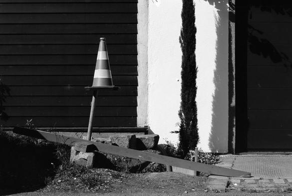 The black and white image shows a traffic cone balanced on a pole, surrounded by construction debris and a wooden beam on the ground. In the background, a building wall is divided into dark horizontal siding and a light plastered section with a tall, narrow tree casting a sharp shadow.