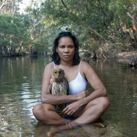 A First Nations woman sits in a river with an owl perched on her arm.