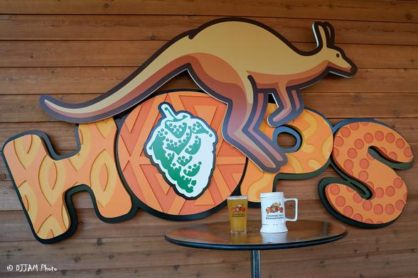 In the image, there is a wooden wall with a large sign that says "hops" in an orange color. On the right side of the sign, there is a coffee cup placed near the top of the sign. The sign is adorned with several cartoon kangaroo pictures, adding a fun and lively element to the scene. The wooden wall provides a rustic backdrop to the scene, creating a visually appealing setting for the coffee cup and the sign.