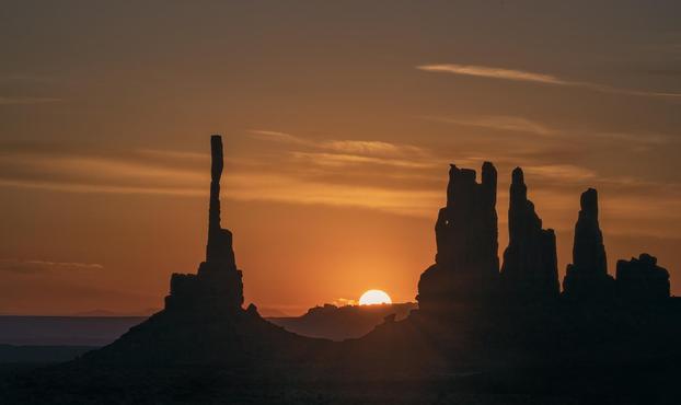 A big yellow sun peeks halfway over the horizon illuminating an orange and gold sky and silhouetting rock formations in Monument Valley.