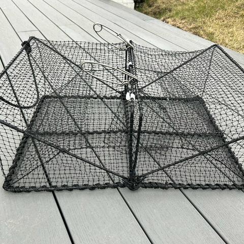 In the image, there is a square-shaped trap with a black mesh exterior, sitting on a gray wooden surface. The trap is open, revealing a metal handle on the front, suggesting that it is intended for use in catching or handling small animals.