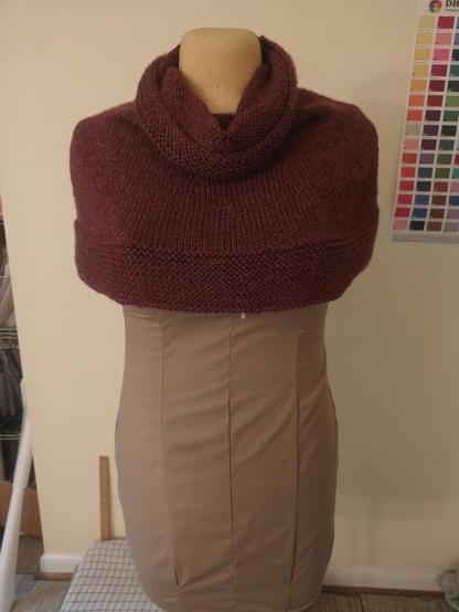 A short, maroon capelet that ends just below the bust with a cowl neck and contrast seed stitch hem.