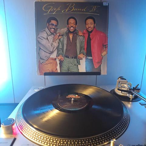 A vinyl record sits on a turntable. Behind the turntable, a vinyl album outer sleeve is displayed. The front cover shows 3 members of the Gap Band looking at the camera while posing together.