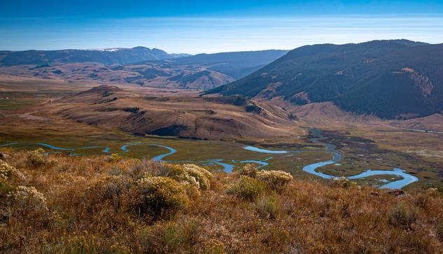 A glassy river winds through a yellow grassy valley with blue hued mountains and a blue sky in the background.