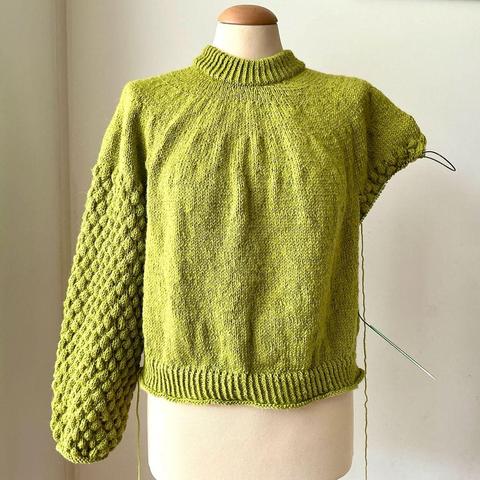 A pear-green knit sweater on a mannequin against a white background. One sleeve is knit in bubble stitch, the other one is still in progress.
