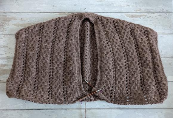 A photo from above of a mauve knitting WIP on a white wooden background. The knitting is the top of the body of a cardigan knit in the round, back and fronts joined but without sleeves yet. It has a textured fabric consisting of honeycomb cable columns divided by lace columns.