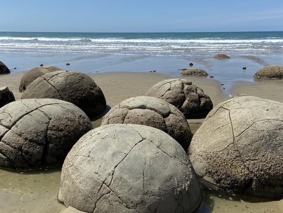 Halb in den Sand eingegraben, sieht man dicht an dicht große, graubraune Gesteinskugeln am Strand liegen. Die Oberflächen sind von feinen Rissen durchzogen, manche scheinen bald aufzubrechen.