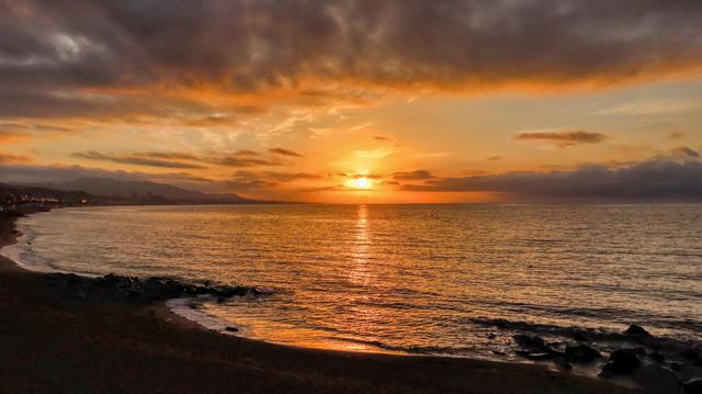 Aquesta imatge captura una posta de sol impressionant sobre l'oceà. El sol, un disc taronja brillant, s'enfonsa per sota de l'horitzó, pintant el cel amb una varietat de tonalitats càlides, des de taronges profundes fins a grocs daurats i roses suaus. Núvols densos i dramàtics es van estendre pel cel, creant un efecte de textura i profunditat.

L'oceà tranquil i serè reflecteix la brillantor del sol ponint, creant un efecte mirall daurat a les seves aigües. La costa, amb les seves roques fosques i sorra fosca, contrasta amb la lluminositat del cel i del mar, creant una escena visualment sorprenent. El litoral, suaument envoltat d'onades, afegeix una sensació de pau i tranquil·litat.