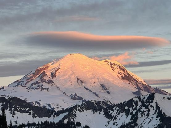 A snow-capped mountain at sunrise, showcasing orange hues on the peak, surrounded by rugged terrain and clouds in the sky.