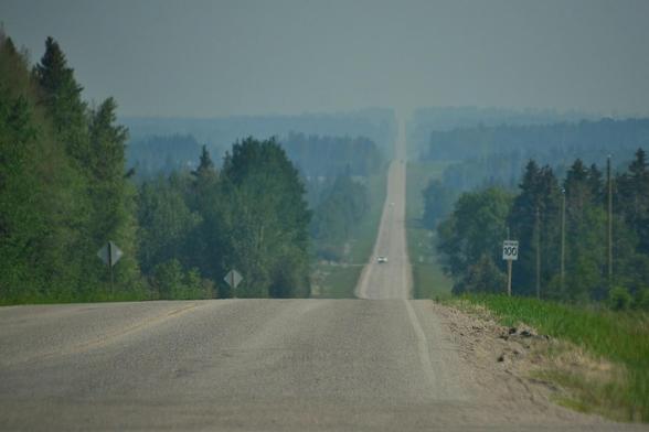 rom the car, looking across a backroad intersection to a paved road that drops dramatically down a hill then continues through a smoke hazed valley, through a couple more hills and dips and rising to a higher distant ridge nearly erased by the smoke. The nearest bit of roadside to right has low fresh grass, then just below the crest of the first hill is a speed limit sign showing 100km/hr. A line of power poles marches down the hill and both sides are lined with dense stands of mixed woods after the mowed ditches,the woodland interrupted every so often by grassy incursions of fields, driveways, side roads.