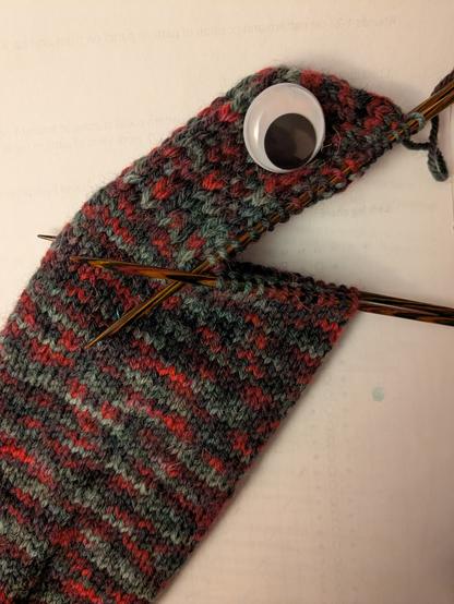Sock in grey and red variegated yarn, with a googly eye on the heel.