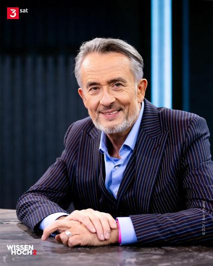 Bild von Moderator Gert Scobel im TV-Studio. Er sitzt am Tisch und lächelt in die Kamera. Unten links zu sehen ist das Logo des Wissenschaftsabends bei 3sat: Wissen hoch 2.