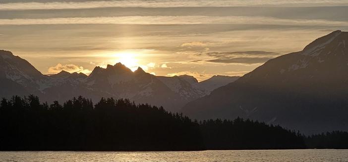 Sunrise over mountains, hazy in the background, you can just see snow at the tops. In the mid-ground there are silhouettes of small islands, with a thin strip of burnished sea at the bottom. Everything is tinged a brassy yellow - looks like an old-fashioned tintype.