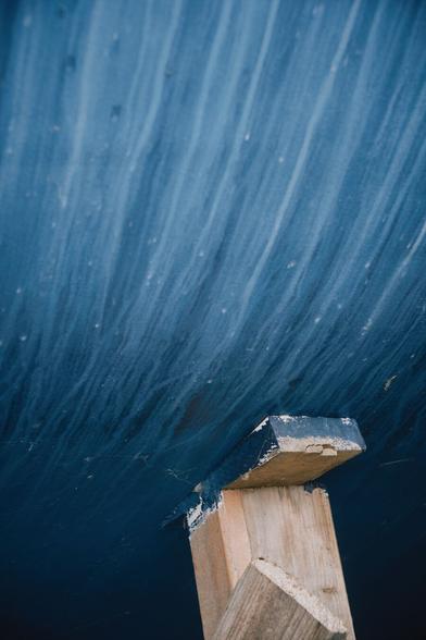 Close-up view of the underwater portion of a ship hull in on-shore maintenance. It curves inwards from the top to the bottom of the photo. The hull is dark blue, but many drops of water have carried white paint from the above water section onto the blue so that there are many layered lines of varying shades of blue running down the hull. In the lower right third the hull is being held by a wooden beam of about an arm‘s thickness.