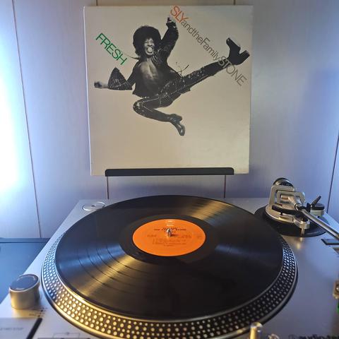 A vinyl record sits on a turntable. Behind the turntable, a vinyl album outer sleeve is displayed. The front cover shows Sly Stone jump kicking through the air.