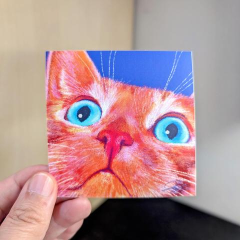 Photo of a left hand holding a square sticker. The sticker is a matte painting of an orange cat with no thoughts.