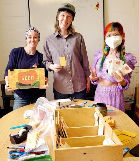 Medium shot of three people standing behind a round, light-colored wooden table. The background is a white wall with a whiteboard containing handwritten text. The table is covered with a wooden organizer box containing seed packets, several unmarked yellow envelopes, a plastic container with pens and scissors, and a tape dispenser. The person on the left wears a patterned headscarf and holds a wooden box with a seed library graphic. The person in the center is wearing a vertically striped shirt and a baseball cap with a logo, holding up a yellow card. The woman on the right (me) has rainbow-colored hair and wears a white mask, giving a peace sign with her left hand and holding up seed packets in the right hand. The overall tone is bright and cheerful.