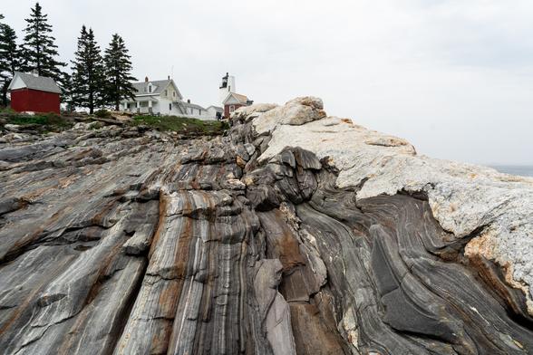 A photograph of a geologically-interesting rocky shoreline with a cylindrical white lighthouse and keeper’s house in the background. The rocks are a grayish metamorphic rock called gneiss that is heavily striated due to its origins as a sedimentary rock with many layers. The stripes in the rock run perpendicular to the angle/location the photo was taken from. The rocks are not only banded but also have a slightly wavy or undulating appearance to them. On the right side of the frame there is a band of whitish-gray igneous rock called pegmatite that runs along top a high point in the rock. All of the lines in the rock look like they are running toward the lighthouse.