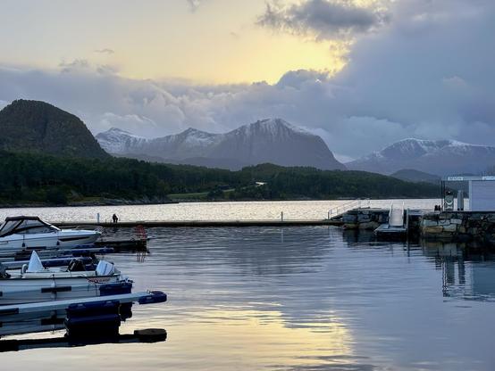 Sunset over a calm lake with moored boats on the left and a dock extending from the right, under a cloudy sky with snow-capped mountains in the background. Two people stand by the water near the center.