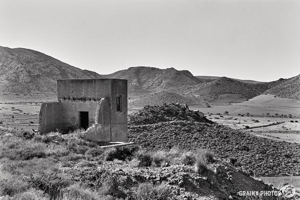 A black-and-white photograph, possibly taken with a Nikon F using Ilford Delta 100 film, captures a deserted, crumbling building overlooking a vast, hilly landscape. The barren and rugged scene with distant mountains and clear sky evokes isolation and timelessness.