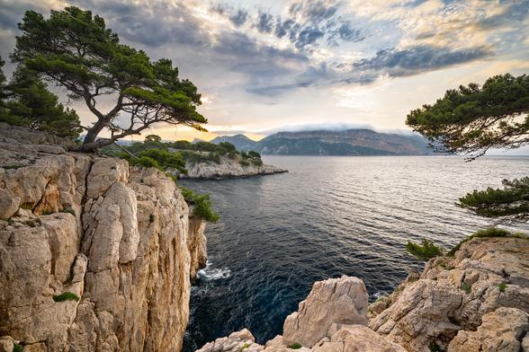 Zu sehen ist eine Meeres-Szenerie bei Sonnenaufgang mit orangenen Granitfelsen im Vordergrund, Meerwasser im Mittelgrund und einer aufgehenden Sonne im Hintergrund, über der die Wolken stehen.
