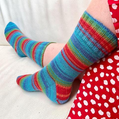 A woman wearing rainbow socks and a red-with-white dotted dress is trying to capture her new hand-knit socks, while sitting on a white leather couch.
