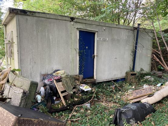 a photo of an abandoned Portakabin site office surrounded by dumped rubbish on the edge of woodland.