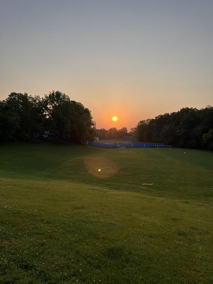 Sunrise over a green grassy school field. There are trees in the background. The sun is a bright yellow-orange ball. There are no clouds in the dark sky.