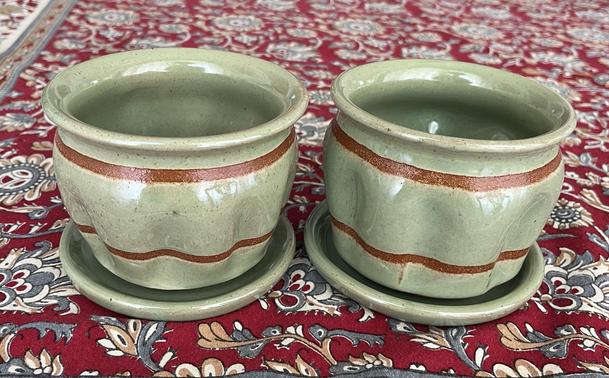 Two planters with matching dishes glazed with Warm Jade. The planters have vertical ridges pushed all the way around them. There are two stripes of raw clay wrapping around the circumference of the pots that were made using tape resist. The pots are sitting on a floral tablecloth, with red being the dominant color.