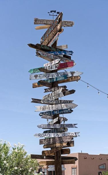 A narrow color portrait photo of a very tall comic sign post with wooden planks pointing in many different directions toward various cities. At the top is a weather vane with a Jeep like vehicle mounted on top of an arrow and below that another metal bar designating the directions "N" for north and "S" for south. 

Among the forty three different signs, not all are legible in the photo, are:
Riff Rat Rancho
Bluhofs
Cabo San Lucas
Virgin Islands
Deset Bar 72mi
Ensanada MX 291mi
Cactus Ridge
Quartzsite
Cibola Lake
Parker, AZ
Maui, HI 2864mi
Cibola Refuge
Hunt Club
Road Ends 7mi
Belton, TX 1215 miles
Black Creek

Behind the sign is a Spanish style modern adobe house. A large green bush with white flowers is in the lower left. The sky is bright blue.