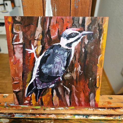 A work in progress 6x6 oil painting on ampersand gessobord.  The woodpecker is gripped onto reddish tree bar and facing its head to the right and looking back at the viewer with one eye.