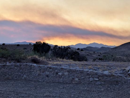 Dark smoke and yellow-orange haze at sunset through the smoke from nearby wild fires. The foreground is a rock wall around my driveway; hills and juniper trees in the middle ground and the Black Range on the horizon. The new fires are on the other side of the Black Range.