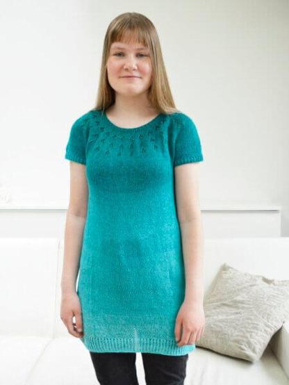 A young woman standing in front of a white wall and couch, wearing a knit tunic with lace detail in the yoke, in a teal to turquoise gradient yarn.