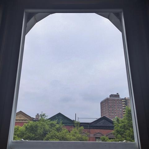 Looking through an arched window thirteen minutes after sunrise the sky is full of grey rain clouds. Pointed roofs of Harlem brownstones with red brickwork are across the street, and a taller apartment building can be seen in the distance. The green tops of two trees are on the bottom and right.