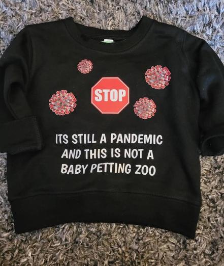 A black long-sleeved sweatshirt is on a gray shag rug. The sweatshirt has a red stop sign image and three images of viruses. Below the images are the words "ITS STILL A PANDEMIC AND THIS IS NOT A BABY PETTING ZOO" in white text.