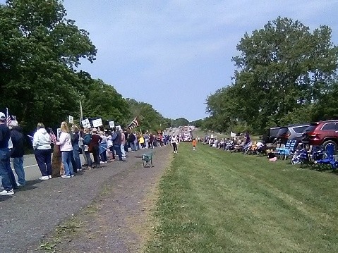 A photo showing a portion of the crowd at the Irondequoit NY No Kings rally. It is taken from behind the crowd, and shows the backs of people lined up along the road, as though watching a parade. Some hold up signs, others wave U.S. flags. The line extends into the distance in this photo. The actual line extended in both directions for a distance of about a mile.