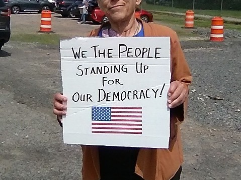 A picture of myself holding my sign at the Irondequoit NY No Kings rally on June 14 2025. The sign says "We the People Standing Up for Our Democracy", with a picture of the U.S. flag. The picture cuts off most of my face, but shows my hands holding the sign, and some brown sleeves. In the background is a gravel area with orange construction barrels.