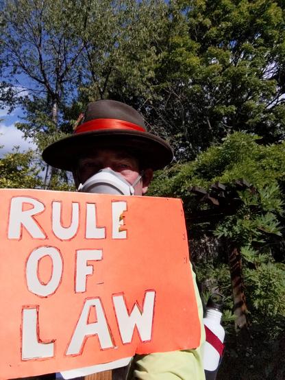 Selfie in hat and Flo mask holding an orange sign with light tan letters, RULE OF LAW.