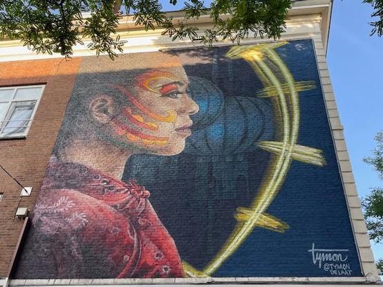 A photo of street art in Rotterdam. Artist Tymon de Laat created this mural called Zodiac for the Chinese community in the city. The mural shows a Chinese woman with decorations on her face. She watches the West-Kruiskade where the Chinese community traditionally celebrates Chinese New Year every year with dragon dances and music and food. 
The woman is wearing an embroidered red jipao. The red-yellow decorations on her face are representing a dragon. In the dark blue background there are Chinese lanterns visible in a lighter blue. There’s a yellow string light (?) in front of the woman. In the right bottom of the mural Tymon’s signature is visible. The sky is blue, the sun is shining a bit on the mural. On the right side there’s a bit of the blue sky visible and parts of a tree. On the left side there’s part of a town house and window visible. On the top side there’s the roof of the building and parts of a tree.